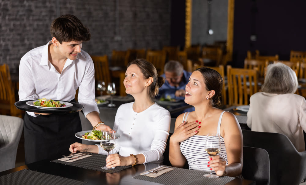 Young waiter serving ordered dishes to women in restaurant