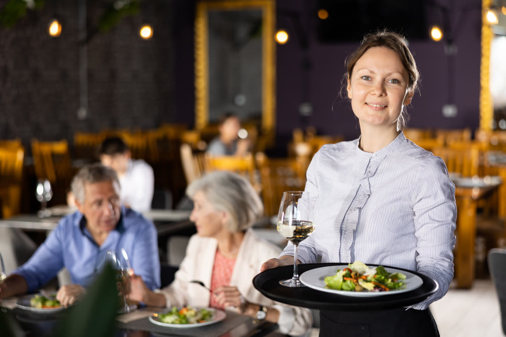 Young female waitress stands with order tray in spacious guest room in restaurant.