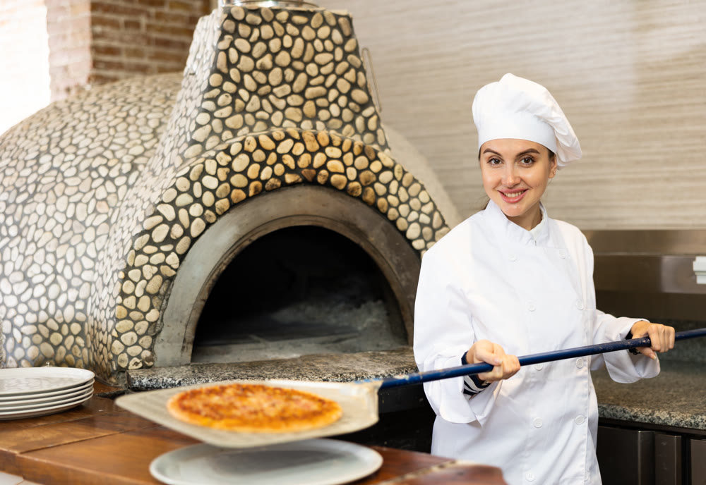 Portrait of young woman chef working in restaurant kitchen, getting ready pizza out of oven