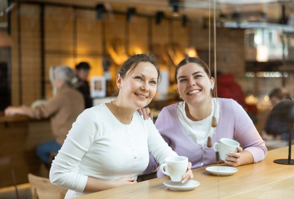 Positive young and middle-aged women in casual wear sitting at table and enjoying coffee or tea in cozy cafeteria