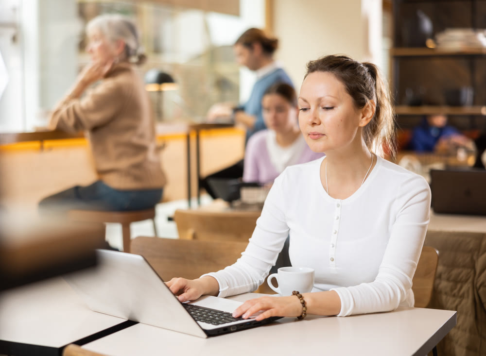 Positive European businesswoman drinking and enjoying coffee while working on laptop in cafe. Freelance and remote work