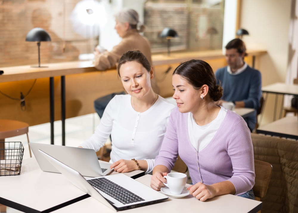 Portrait of two positive young females surfing Internet on notebook while drinking tea or coffee in cafeteria