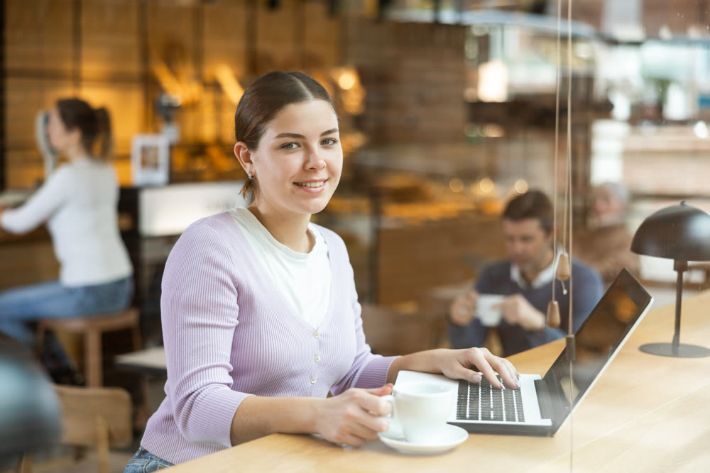 Young millennial woman sitting at table, using laptop and enjoing cup of tea in coffee house. Modern female lifestyle concept