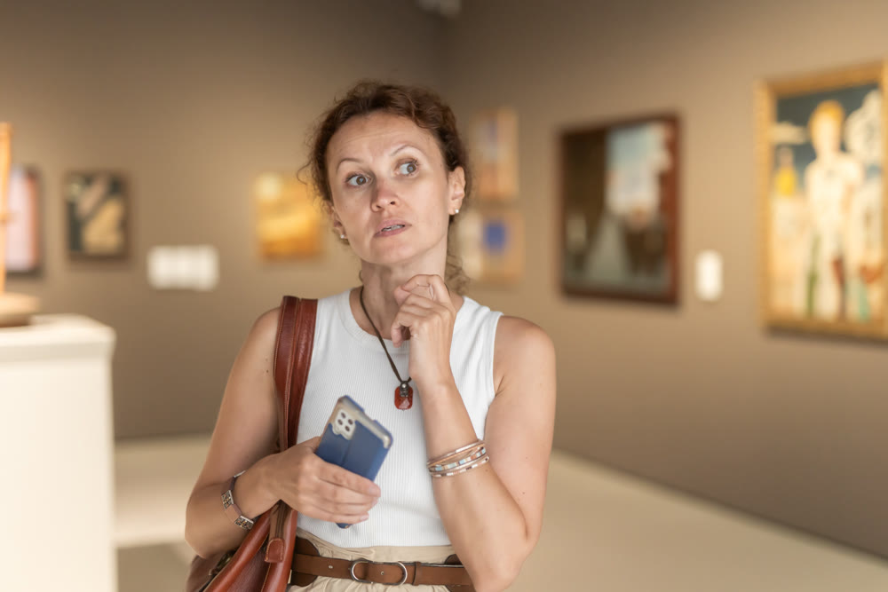 Concentration woman with smartphone in hands thoughtfully admiring exhibition of works of art during sightseeing tour