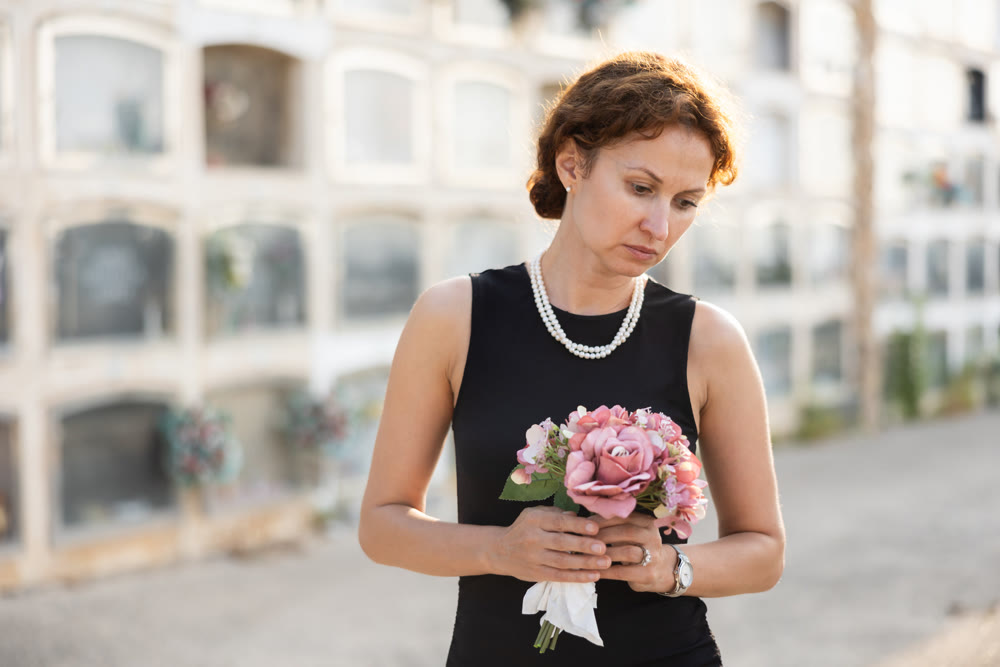 Sad woman in black dress with bouquet of flowers and handkerchief in hands, stands in cemetery