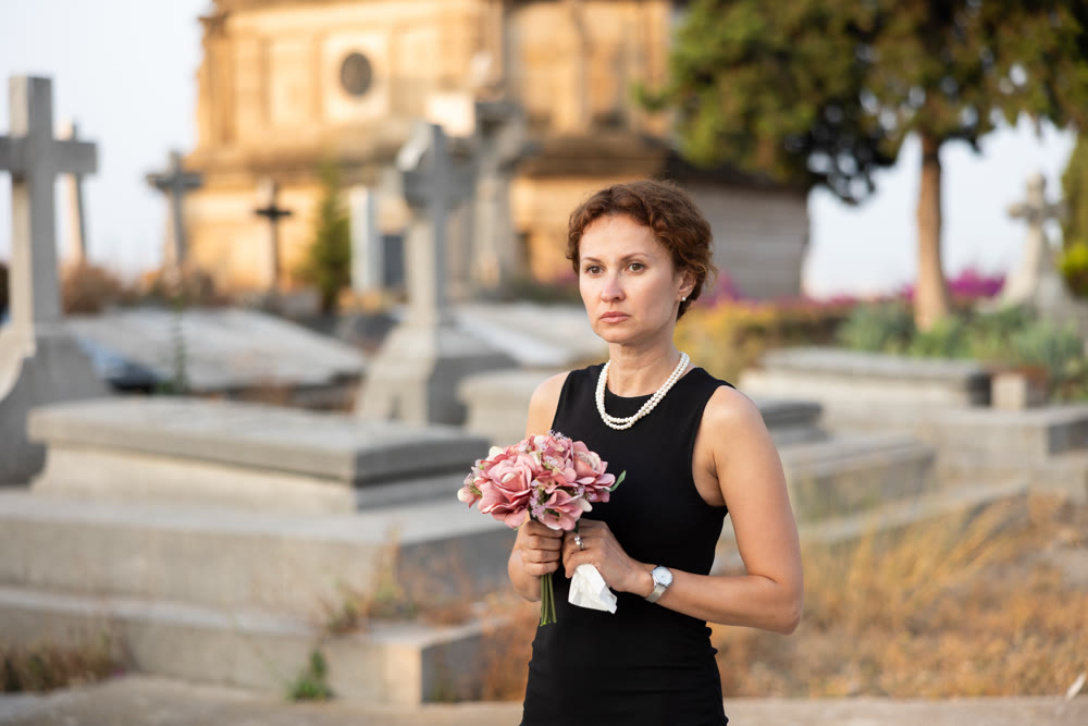 Sad woman in black dress with bouquet of flowers and handkerchief in hands, stands in cemetery