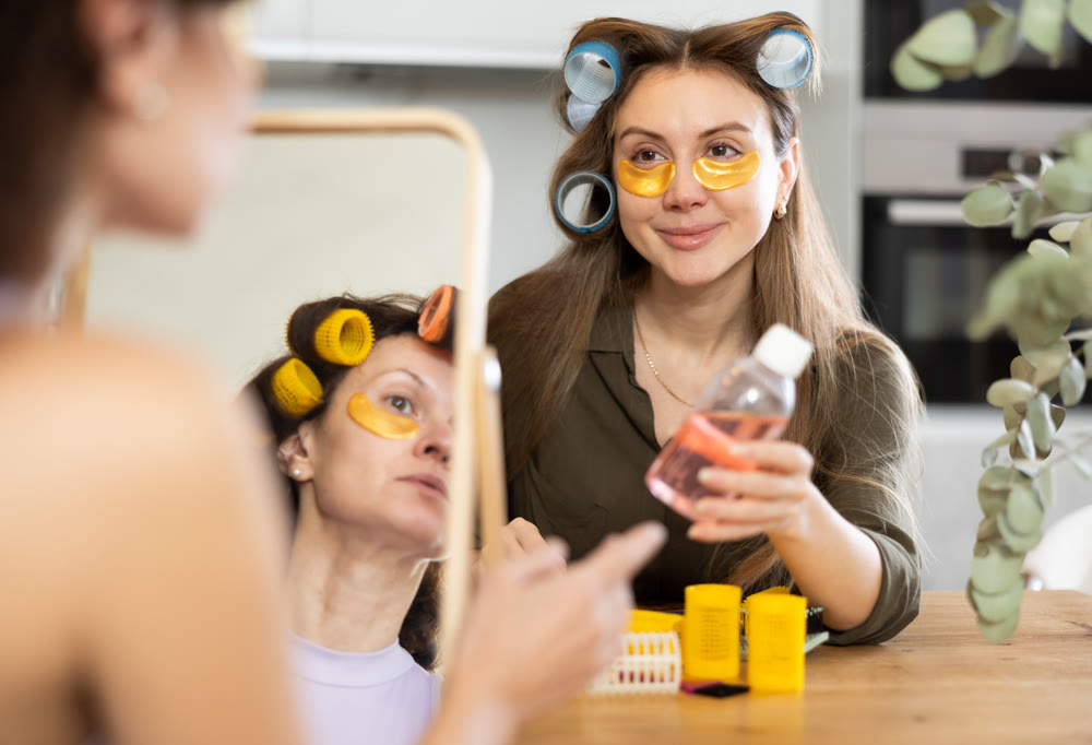 Woman in hair rollers applying eye patches in front of mirror with friend