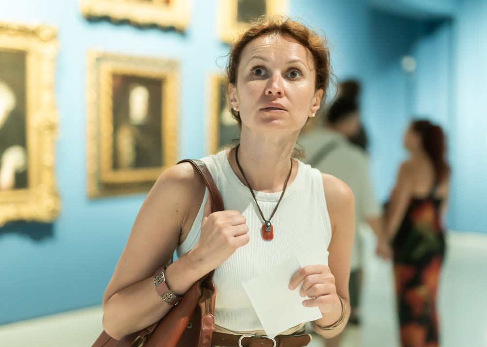 Interested woman looking closely at paintings in art gallery and holding tour sheet in hands