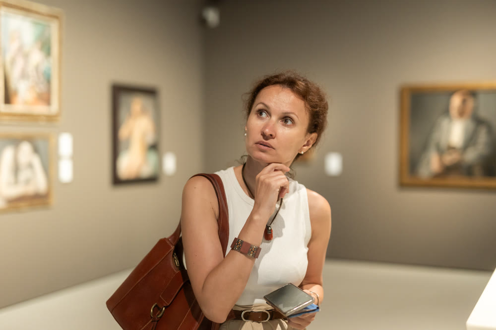 Attractive young woman in casual clothing with phone in hands looking painting in museum