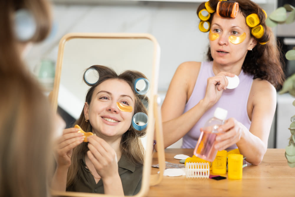 Woman in colorful curlers applying under eye patches by mirror with bestie