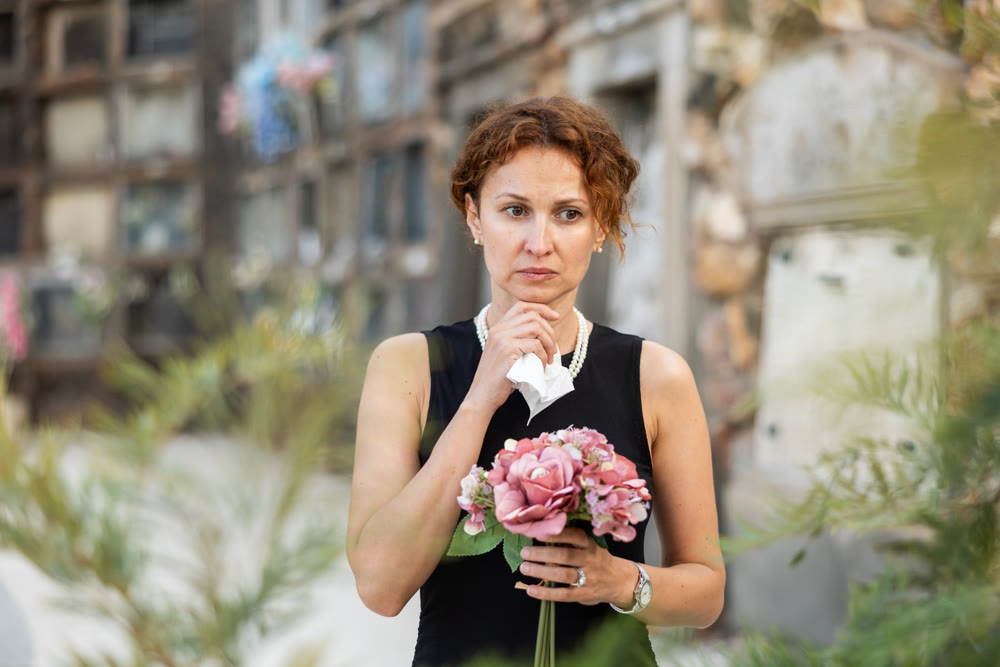 Sad woman in black dress with bouquet of flowers and handkerchief in hands, stands in cemetery