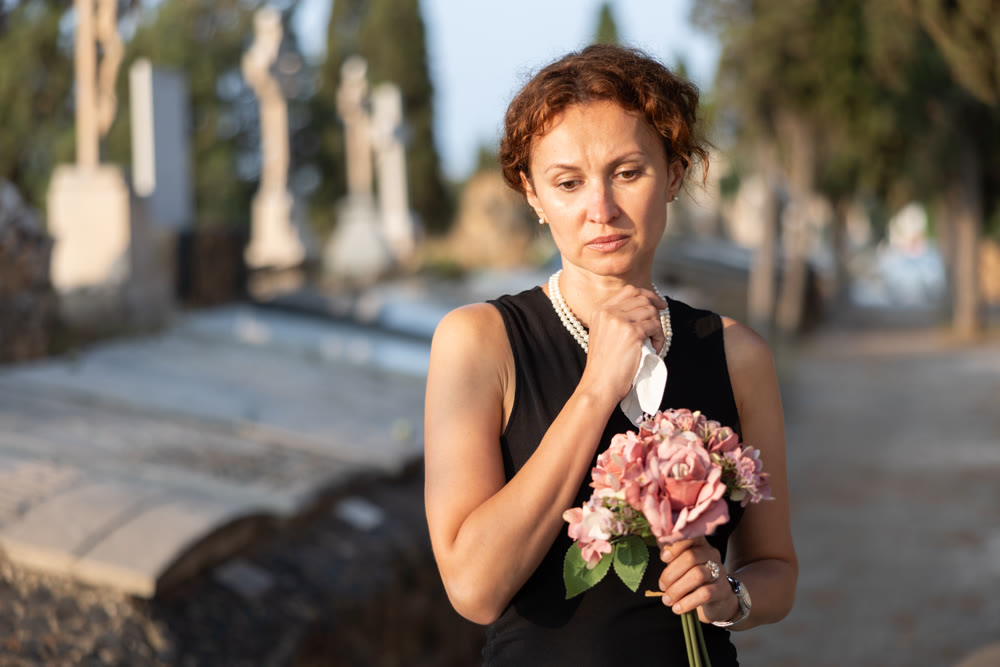 Sad woman in black dress with bouquet of flowers and handkerchief in hands, stands in cemetery