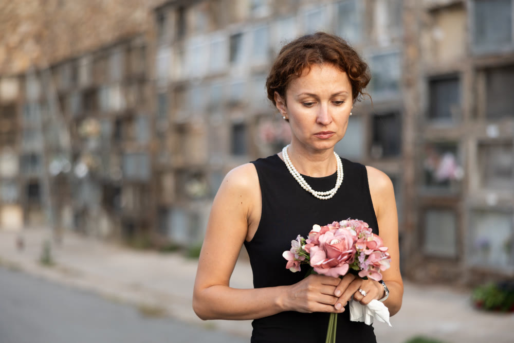 Sorrowful woman in black standing in cemetery with flowers