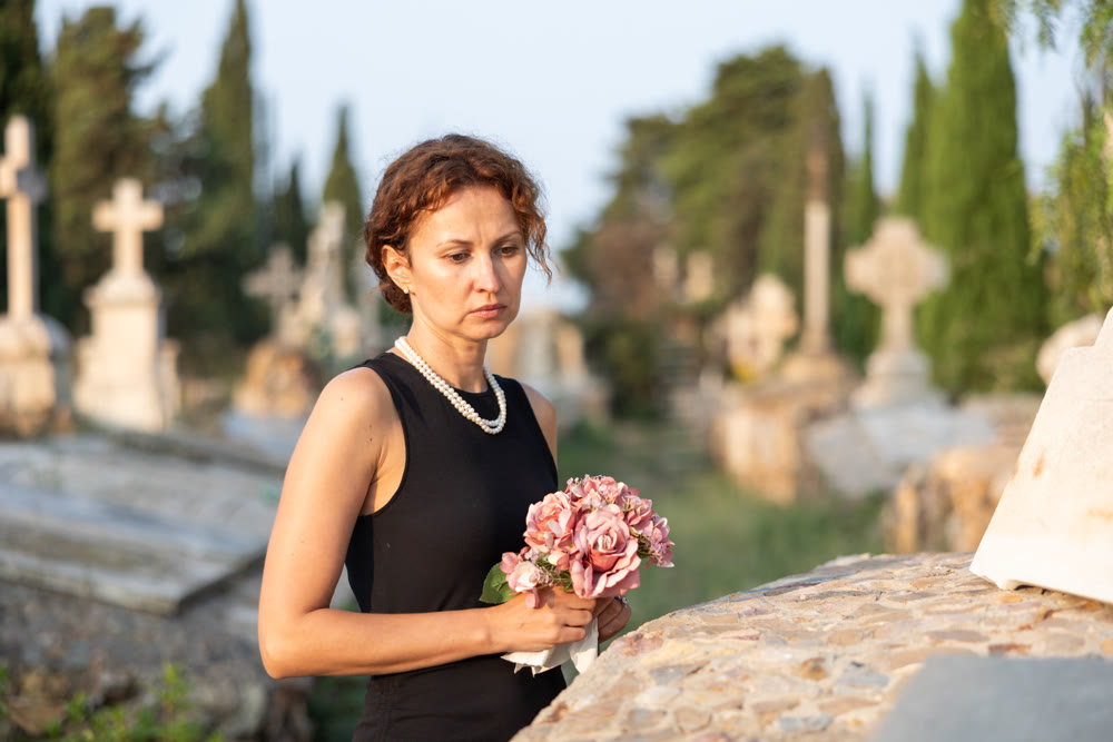 Sad woman in black dress with bouquet of flowers and handkerchief in hands, stands in cemetery
