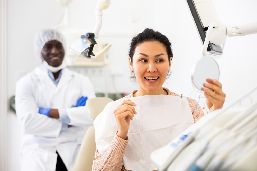 Woman satisfied with result of teeth treatment looking in mirror at dental office