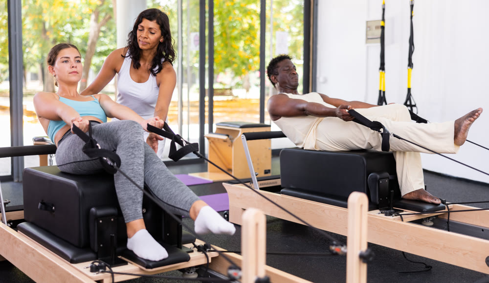 Latin female instructor helping positive girl practicing pilates on reformer