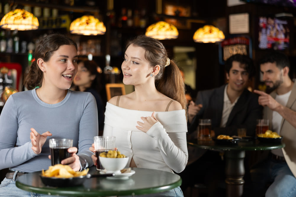 Two women chatting in a bar with guys in the background