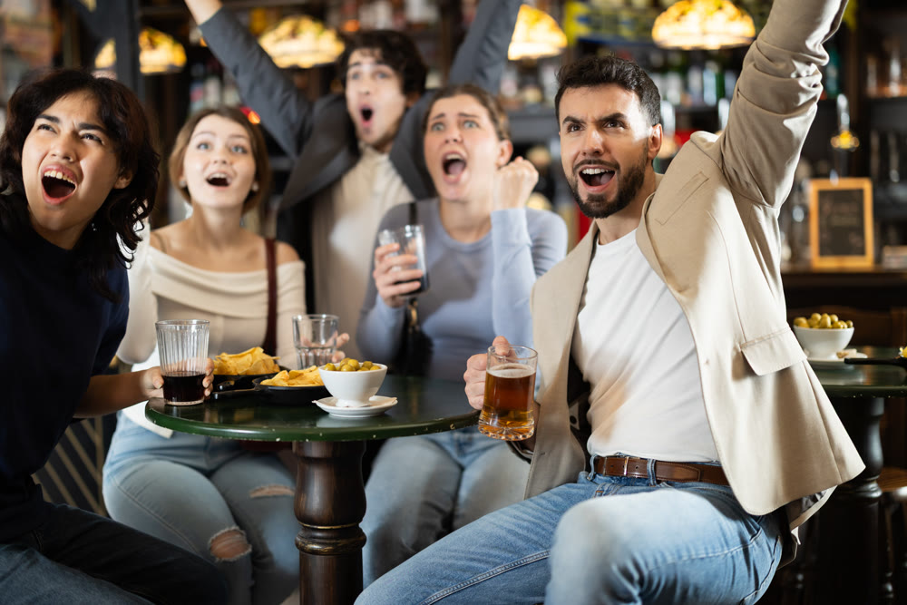 Fans watching match at a sports bar, cheering with drinks, energetic atmosphere, friendship and fun, people enjoying game together indoors