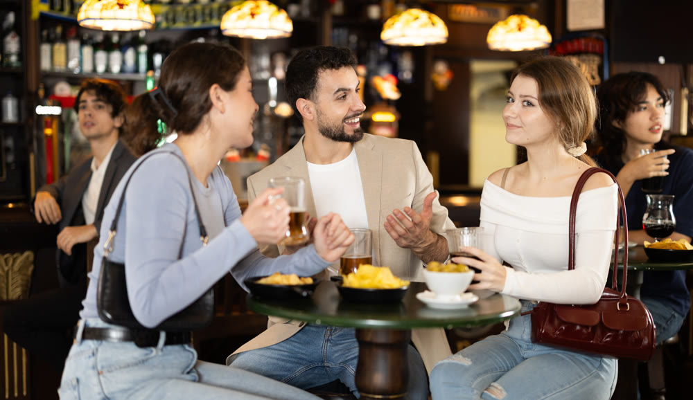 Two women actively chatting with a guy in a bar with a glass of beer