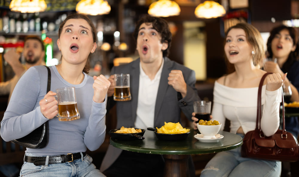 Group of emotive young friends supporters cheering for favorite team celebrating victory with glasses of alcohol in beer pub