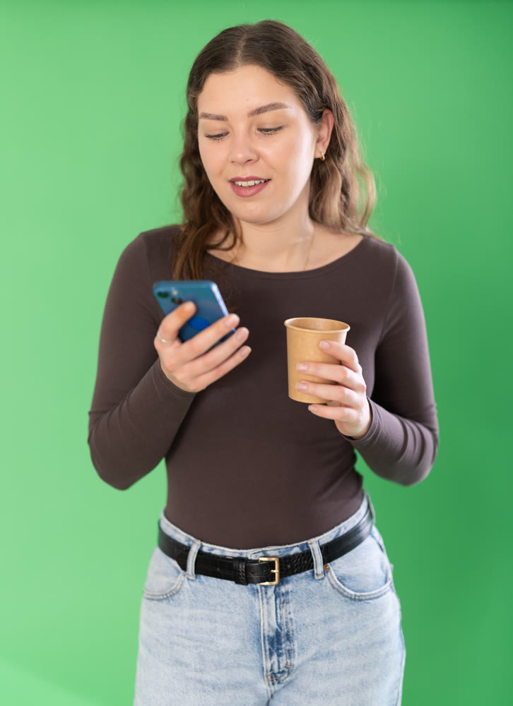 Young woman scrolling smartphone and drinking coffee in front of green screen