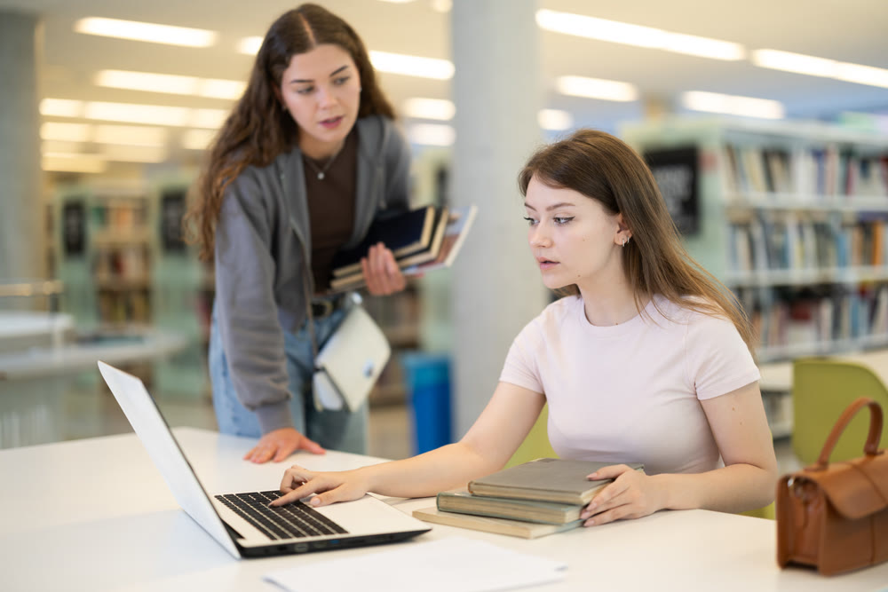 Two girls studying for an exam together in university library using books and laptop