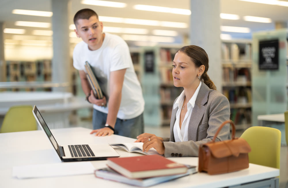 Girl and boy studying for exam together in library - using books and laptop