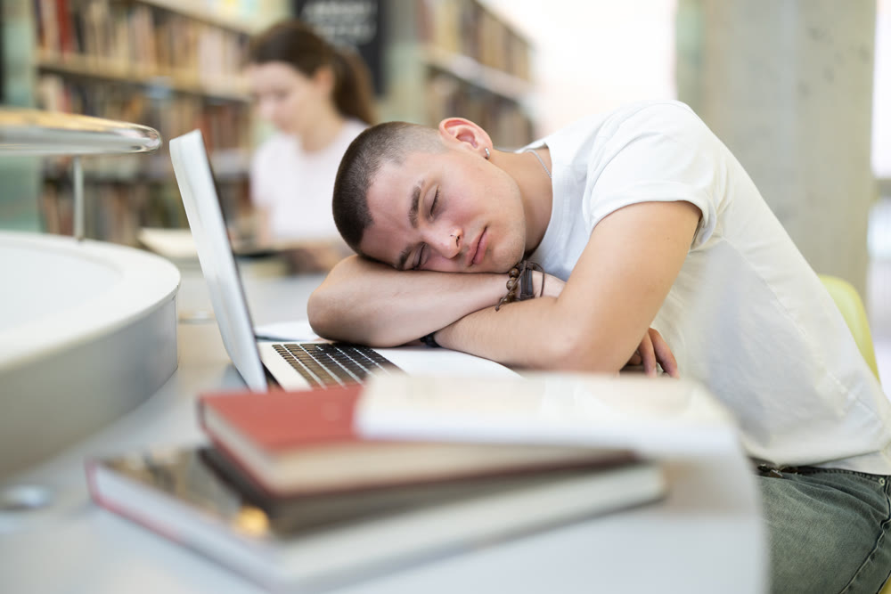 Tired student fell asleep at table next to a laptop and books in public library