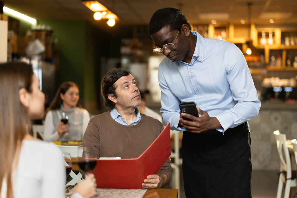 African male waiter communicates with man customer, serve couple in restaurant