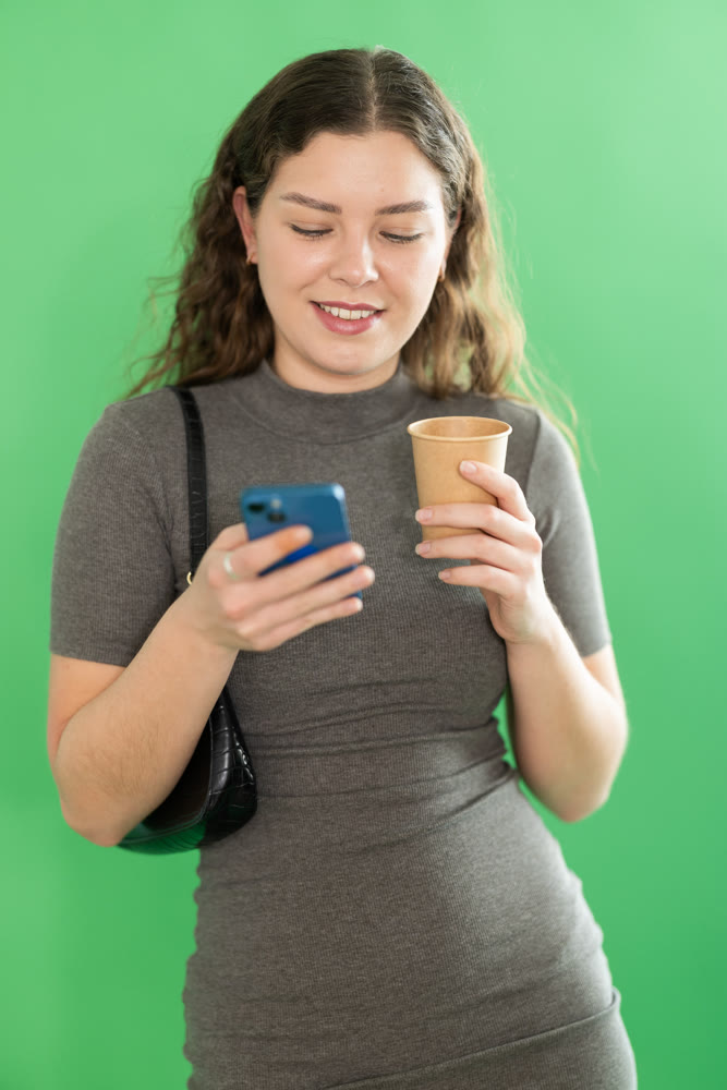 Pretty young woman scrolling through her smartphone screen against and drinking coffee green screen background. Chroma key compositing