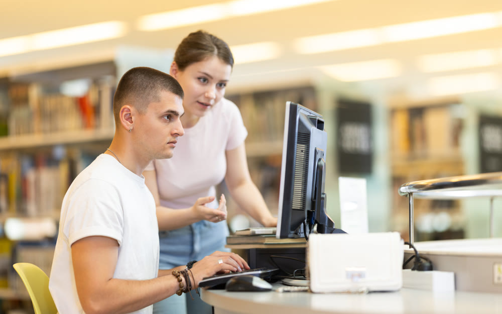 Male student works on computer in library and talks with female friend.