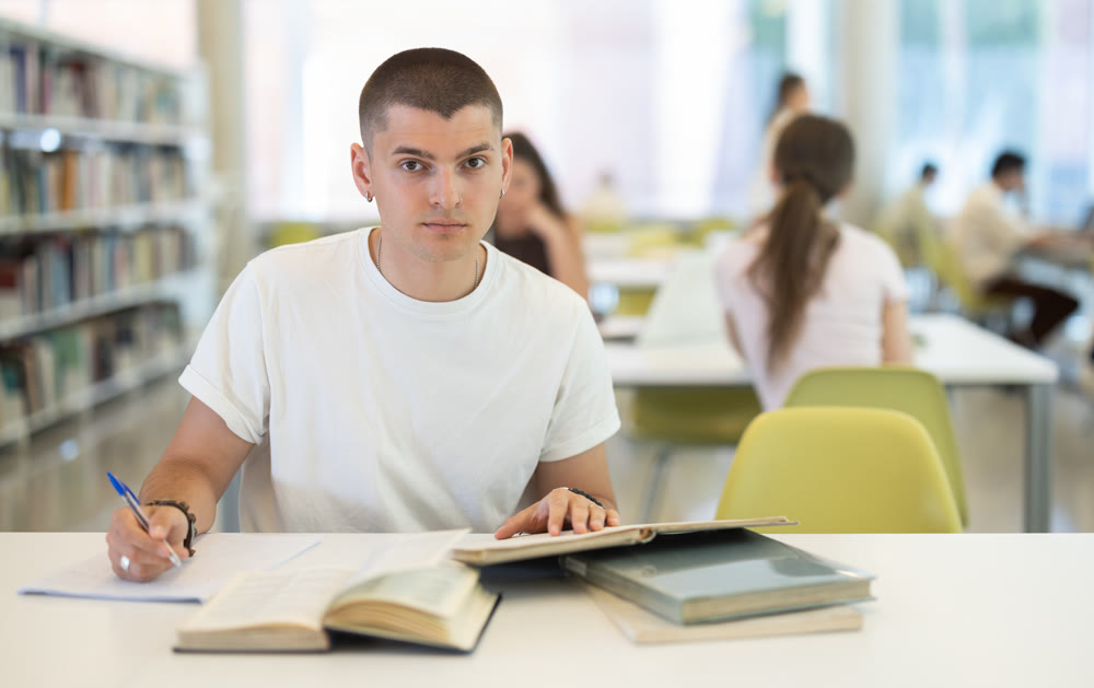 Male student sits at table in library and studies, reads books and takes notes