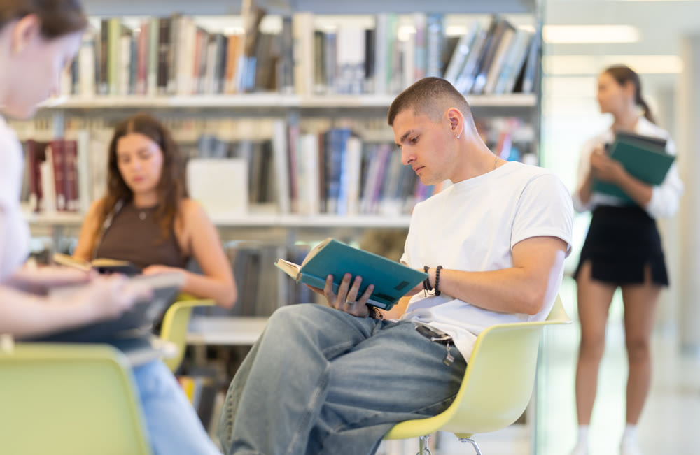 Male student reading book on chair in library