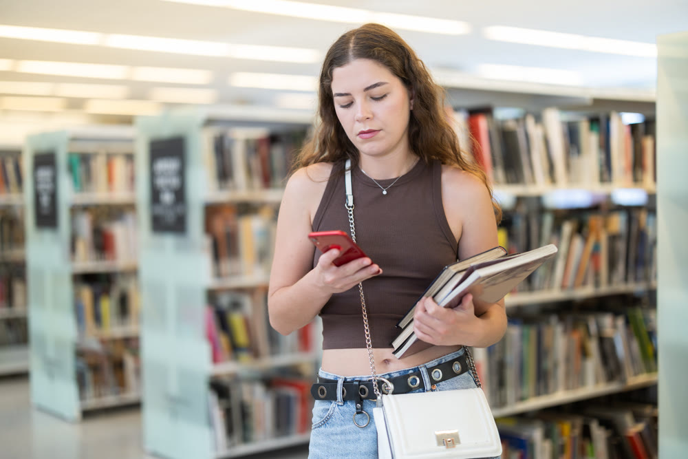 Female student stands in library with stack of books and scrolling though phone screen