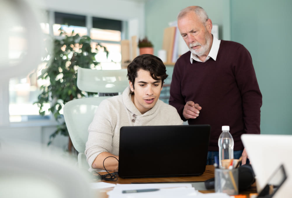 Elderly man helping young male colleague work on laptop