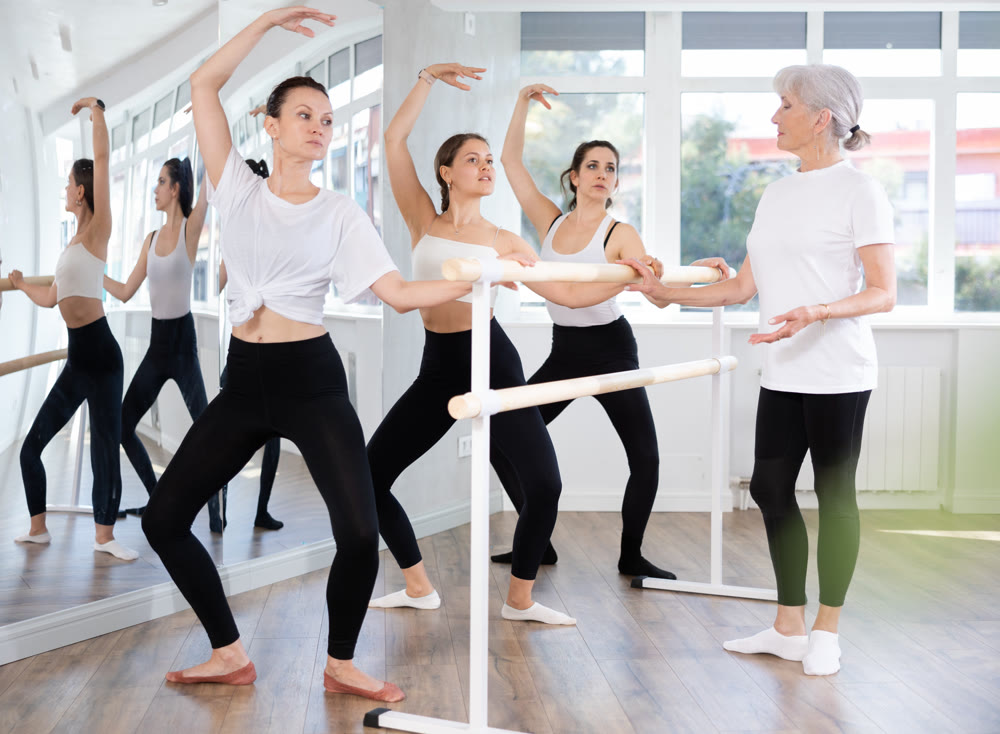 Teacher conducts class with girls in ballet studio, observes doing grand plie exercise.