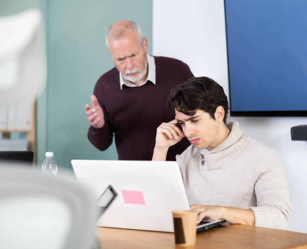 Elderly man scolding young male colleague working on laptop