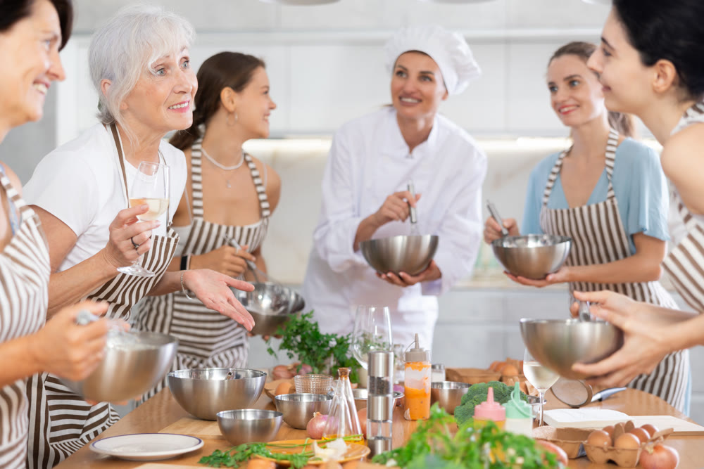 Aged woman holding wine and talking to other cooking course members whipping souce in bowls