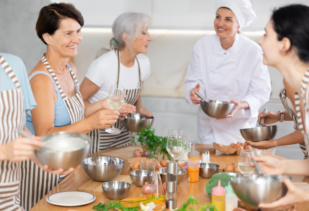 Aged woman holding wine and talking to other cooking course members whipping souce in bowls