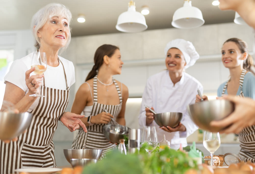 Aged woman holding wine and talking to other cooking course members whipping souce in bowls