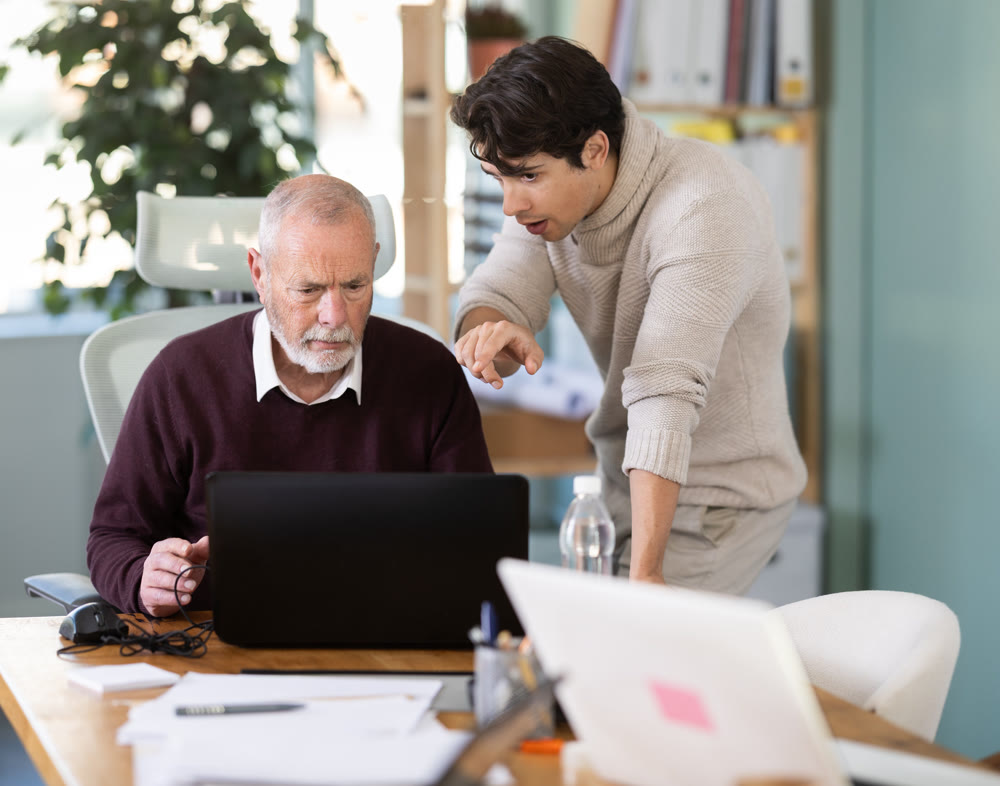 Young man helping elderly male colleague work on laptop