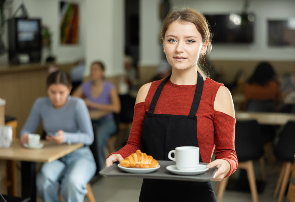Polite waitress holding tray with coffee and croissant in cafeteria
