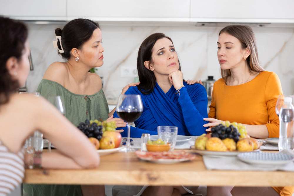 Sad Caucasian woman sits silently in kitchen, female friends sympathize and comfort woman