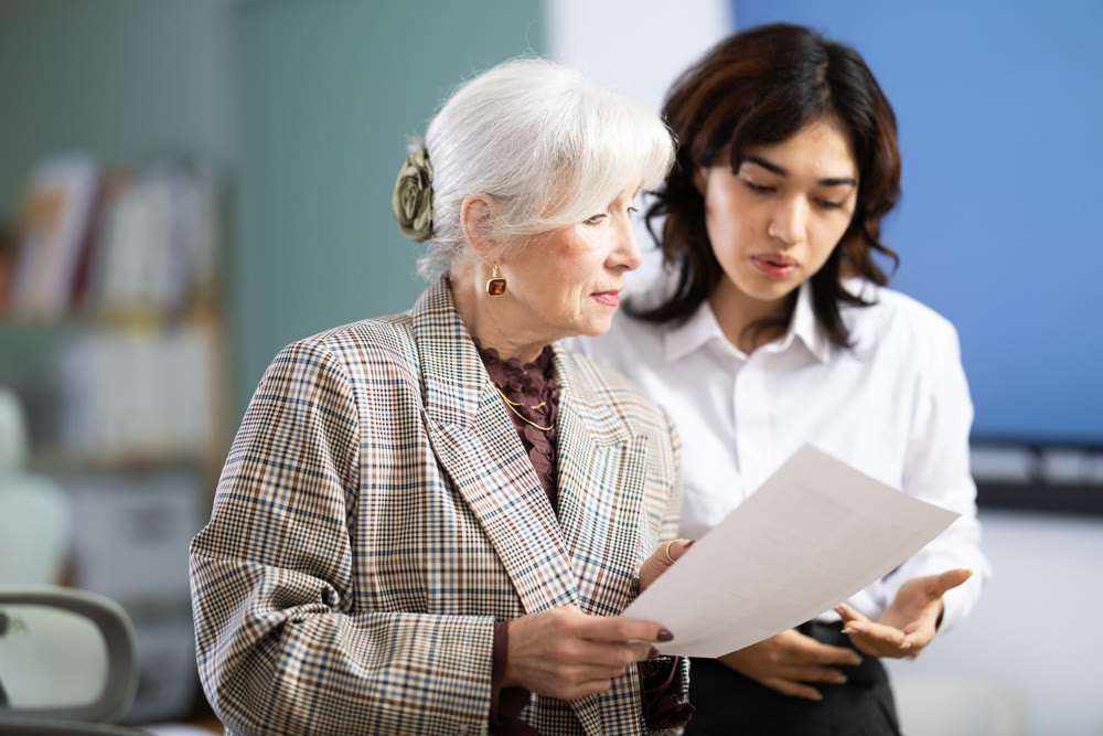 Mature woman and young colleague discussing documents while standing in office