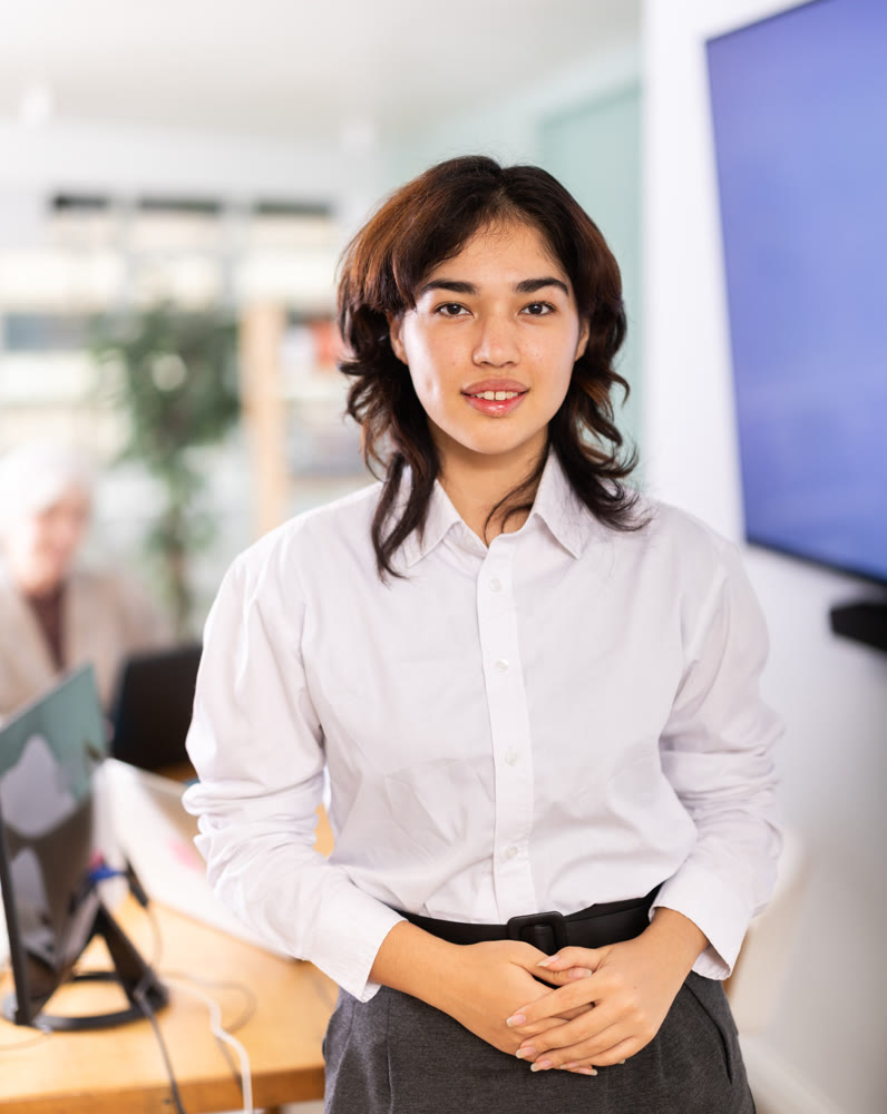 Asian girl employee stands in buzzing office, in middle of working day