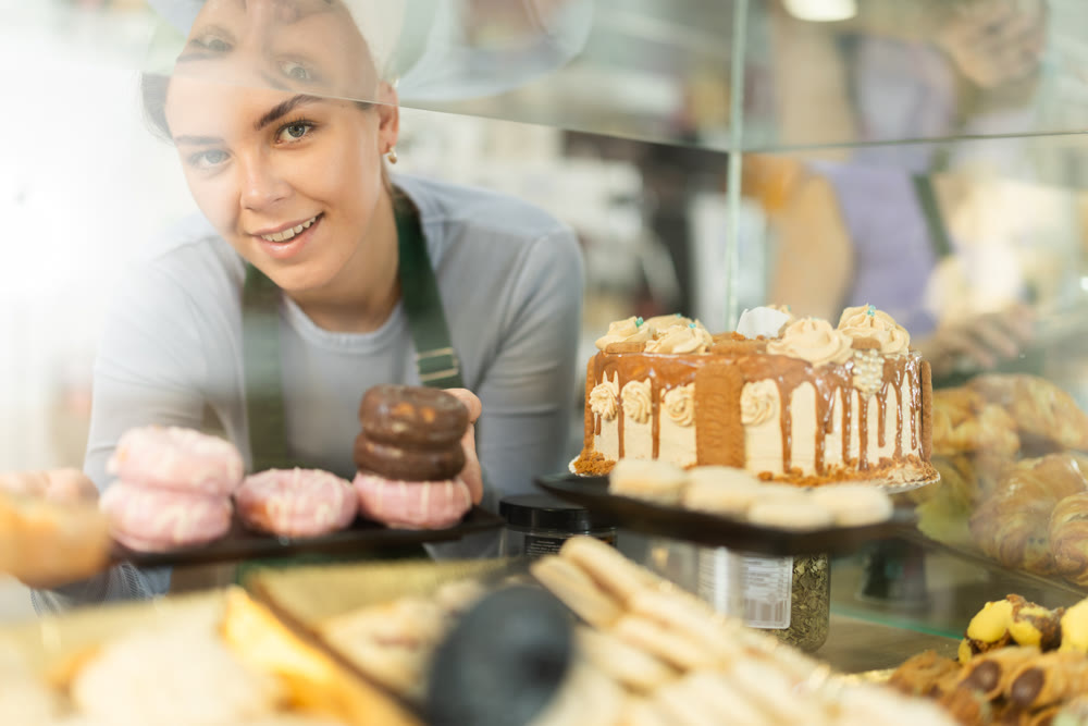 Positive young girl offering doughnuts standing behind glass display
