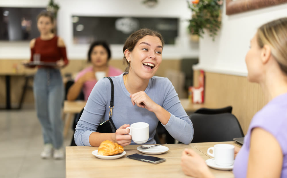 Young girl chatting with friend over coffee and croissant in cafeteria