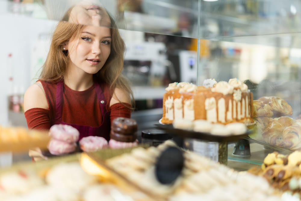 Positive young girl offering doughnuts standing behind glass display