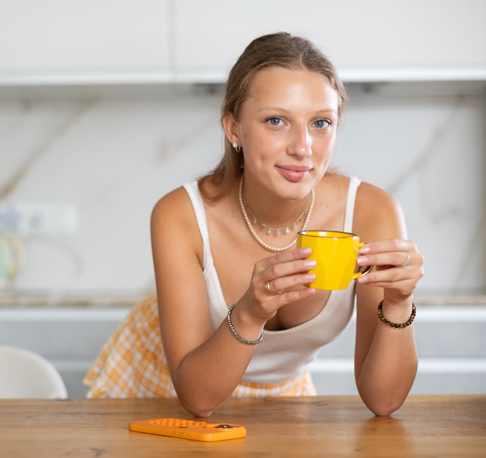 Attractive young girl wearing home jersey and short summer shorts enjoying cozy morning on kitchen with cup of coffee