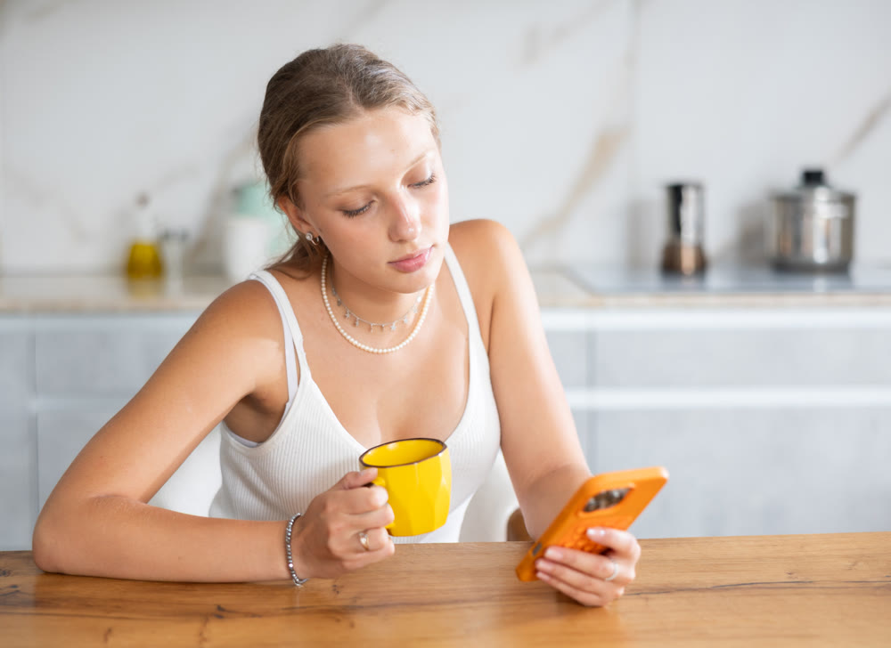 Relaxed young girl looking at smartphone while drinking coffee in kitchen